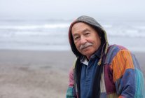 Seitenansicht eines älteren Mannes im Mantel, der am abgelegenen, leeren Strand der Meeresküste steht und in die Kamera blickt — Stockfoto