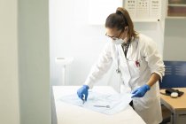 Doctora joven en uniforme y máscara médica colocando equipo de cirujano en la servilleta en la habitación - foto de stock