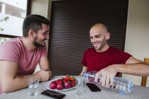 Pareja gay comiendo fresas y bebiendo agua en la mesa en la cocina - foto de stock
