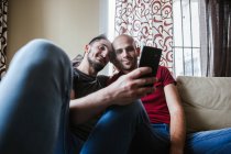 Feliz pareja gay relajarse en el sofá y compartir el teléfono inteligente - foto de stock