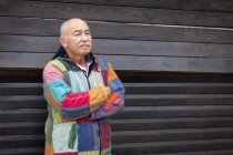 Hombre mayor reflexivo usando chaqueta colorida simple y de pie con los brazos cruzados contra la pared de madera de la casa - foto de stock