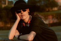 Nachdenkliche Frau mit trendiger Sonnenbrille und schwarzem Hut, die sich im Sonnenlicht an die Hand lehnt — Stockfoto