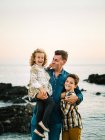 Hombre de mediana edad con sus hijos en la orilla del mar sonriendo y abrazándose - foto de stock