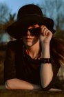 Пряма жінка в модних сонцезахисних окулярах з чорним капелюхом, що спирається на руку і дивиться на камеру на сонячному світлі — стокове фото