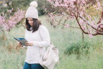 Reisende mit Rucksack liest Reiseführer, während sie in der Frühlingslandschaft in der Nähe blühender Bäume spaziert — Stockfoto