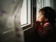 Piccola ragazza sbirciando attraverso la finestra a casa — Foto stock