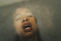 Femme noire sensuelle avec les yeux fermés gémissant et respirant lourdement tout en ayant des relations sexuelles derrière la fenêtre mouillée — Photo de stock