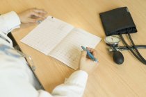 Крупним планом жінка-лікар в уніформі пише на папері, сидячи за столом з тонометром — стокове фото