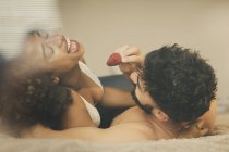 Barbudo chico alimentación alegre novia con fresa fresca mientras está acostado en cómoda cama juntos - foto de stock