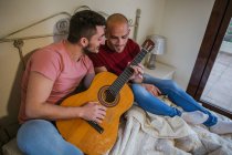 Alegre gay pareja jugando guitarra en dormitorio - foto de stock