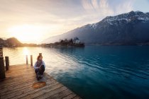 Женщина, сидящая на деревянном пирсе над бирюзовым озером в снежных горах Швейцарии — стоковое фото