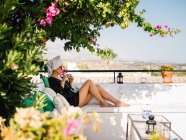 Femme en satin noir robe de chambre et serviette dans la tête reposant sur un balcon bénéficiant de belles vues sur un matin ensoleillé et lumineux — Photo de stock