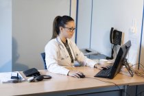 Jeune femme médecin en uniforme naviguant sur ordinateur et assis à table avec tonomètre dans la chambre — Photo de stock