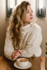 Donna sensuale in elegante maglione a maglia guardando altrove e mescolando caffè aromatico mentre seduto al tavolo del caffè — Foto stock