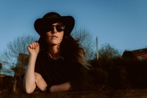 Donna pensierosa che indossa occhiali da sole alla moda con cappello nero appoggiato a portata di mano alla luce del sole — Foto stock
