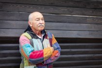 Задумчивый пожилой человек в простой красочной куртке и стоя с руками, скрещенными к деревянной стене дома — стоковое фото