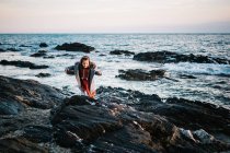 Cute little girl in winter clothes standing on a rocky beach — Stock Photo