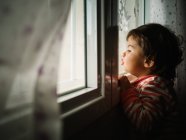 Petite fille regardant par la fenêtre à la maison — Photo de stock