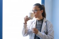 Молода жінка-лікар в уніформі пиття з чашки в кімнаті — стокове фото