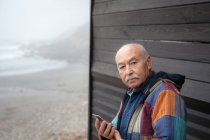Выдержанный серьезный мужчина в куртке, держащий смартфон, глядя в камеру на деревянный дом на удаленном побережье — стоковое фото