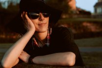 Pensive woman wearing trendy sunglasses with black hat leaning on hand in sunlight — Stock Photo