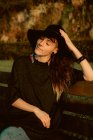 Joven morena sensual con sombrero negro y accesorios sentados en el banco a la luz del sol - foto de stock