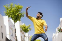 Cheerful bearded male in headphones screaming and taking selfie while standing on city street and listening to music on sunny day — Stock Photo