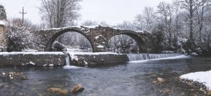 Río que fluye en la nieve bosque de invierno con viejo puente en ruinas - foto de stock