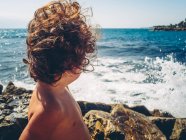 Вид ззаду на анонімну маленьку дівчинку, яка дивиться на море на скелястому пляжі — стокове фото