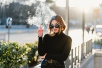 Стильна жінка палить сигарету біля станції метро на сонячній вулиці — стокове фото