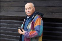 Alter ernster Mann in Jacke hält Smartphone in der Hand, während er auf Kamera gegen Holzhaus blickt — Stockfoto