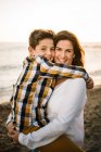Женщина средних лет с сыном на берегу моря, улыбающимся и обнимающим друг друга — стоковое фото