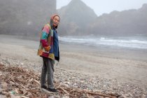 Вид сбоку на старшеклассника в пальто, стоящего на удаленном пустом пляже побережья океана, смотрящего в камеру — стоковое фото