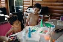 Dos chicos afroamericanos sin camisa usando pintura brillante para hacer cuadros abstractos en la mesa en casa - foto de stock