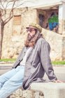 Élégant homme barbu avec les cheveux longs assis sur la rue avec des lunettes de soleil — Photo de stock