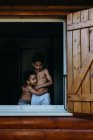 Deux frères afro-américains torse nu regardant par la fenêtre ouverte de la maison en bois tout en s'amusant à la maison ensemble — Photo de stock