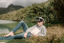 Adolescente joven con gafas de realidad virtual tumbado en la hierba al aire libre cerca de un lago con un libro - foto de stock