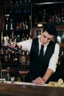 Jovem barman elegante trabalhando atrás de um balcão bar derramando bebida de agitador para um copo — Fotografia de Stock