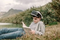 Молодий підліток з окулярами віртуальної реальності лежить на траві на відкритому повітрі біля озера з книгою — стокове фото