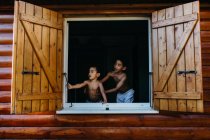 Two shirtless African American brothers looking out open window of wooden house while having fun at home together — Stock Photo