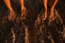 Pernas descalças de mulheres irreconhecíveis em pé na costa molhada perto do mar salpicando à noite — Fotografia de Stock