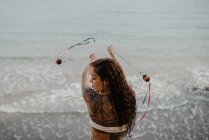 Visão traseira da jovem fêmea tatuada em roupa de banho balançando fios com bolas enquanto dança perto do mar tempestuoso — Fotografia de Stock
