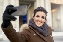 Joven mujer alegre en ropa de invierno tomando una selfie con teléfono inteligente al aire libre en Milán Italia - foto de stock