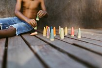 Unerkennbarer, hemdloser afroamerikanischer Junge sitzt auf einer Holzfläche und spielt an sonnigen Tagen mit bunten Zapfen — Stockfoto