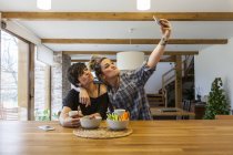 Дві красиві і молоді жінки снідають вдома і розважаються, роблять автофотографію зі смартфоном — стокове фото