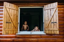 Zwei hemdlose afrikanisch-amerikanische Brüder blicken aus dem offenen Fenster eines Holzhauses, während sie zu Hause zusammen Spaß haben — Stockfoto