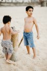 Два афроамериканських брати з палицями бавляться на піщаному березі біля моря. — стокове фото