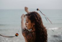 Вид сзади на молодую татуированную женщину в купальниках, размахивающую нитками с мячами во время танцев у бурного моря — стоковое фото