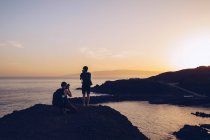 Вид на зворотній бік мандрівників, що стоять на вершині скелі і фотографують прекрасний захід сонця над узбережжям — стокове фото