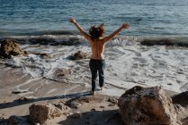 Задний вид босиком топлесс женщина подняла руки шагая в сторону пенных волн бурного моря в солнечный день на природе — стоковое фото